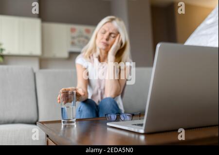 Sad or sick caucasian middle aged defocused female freelancer, suffering from headache, tired of working overtime online, sitting at home in the living room, with closed eyes, need a rest Stock Photo