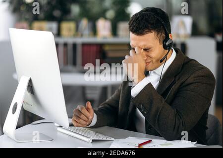 Hispanic male manager or call center operator sits at workplace with headphones, using computer. Tired employee or tutor works without break, have a headache, needing a rest or break Stock Photo
