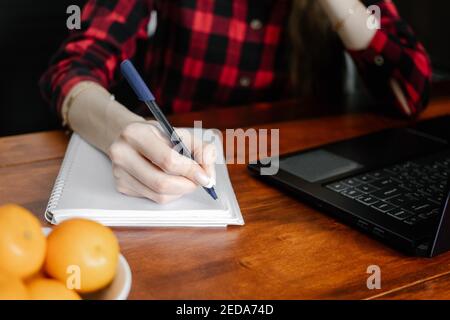 Close up hand of girl college student  writing in workbook sits at desk. Woman learn online on laptop studying from home.