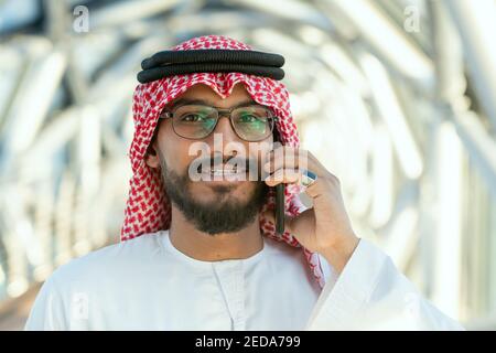 Smiling young Arab delegate or businessman in national attire talking on mobile phone in front of camera in contemporary business center Stock Photo