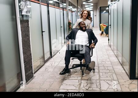 Friendly coworkers take a break from work. Caucasian woman with african american man having fun at the workplace. Multiracial business people in formal wear make a race on office chairs Stock Photo