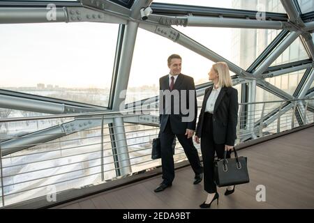 Two foreign mature delegates in elegant suits discussing terms of their partnership while moving along aisle inside modern business center Stock Photo