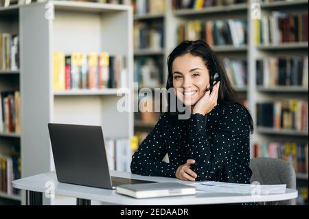 Attractive caucasian young woman, call center worker, student, or successful business woman, in a headset is using a laptop while working online sitting at her work desk and looking at the camera with Stock Photo