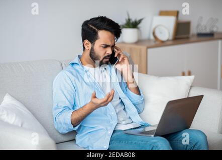 Computer Problems. Frustrated arab guy using cellphone and looking on laptop screen Stock Photo