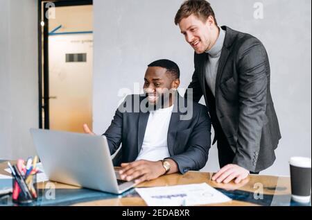 Successful business men using laptop, look at the screen, search ideas for startup. Caucasian male employee gives advice to african american colleague. Young adult coworkers discuss the project, smile Stock Photo