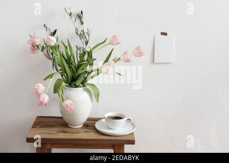 Easter, spring still life scene. Cup of coffee and floral bouquet in ceramic vase on wooden bench. Pink tulips flowers, olive tree branches on table Stock Photo