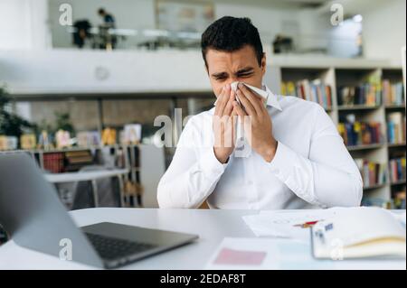 Unhealthy male employee sit at the desk, using a paper tissue, sneezing. Business man feeling unwell, struggling with running nose. Hispanic guy remotely working or studying, while sickness Stock Photo