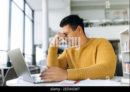 Exhausted male student or employee sitting at the table, massage the eyes, having a headache. Hispanic business man tired from working or studying, feeling unhealthy or stressed Stock Photo