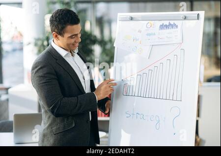 Hispanic smiling man in formal wear, standing in modern office, making a presentation on briefing. Confident male employee shows on marker board a new strategy for company Stock Photo