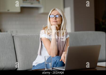 Mature calm caucasian woman with eyeglasses sitting on the couch at home in living room, using laptop, while working remotely, pensive looks away, dreams about weekends or vacation Stock Photo