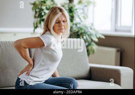 Upset mature woman suffering from backache, unhappy senior blonde sitting on a sofa at living room, feeling discomfort because of pain in back. Stock Photo