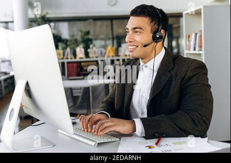 Happy hispanic manager sitting at the desk, wearing headset. Business man work as call center operator, communicate with clients, consulting people. Support service Stock Photo
