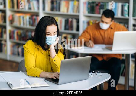 Education during quarantine. Students sit at a distance from each other dressed protective masks on their faces and studying using a laptop Stock Photo