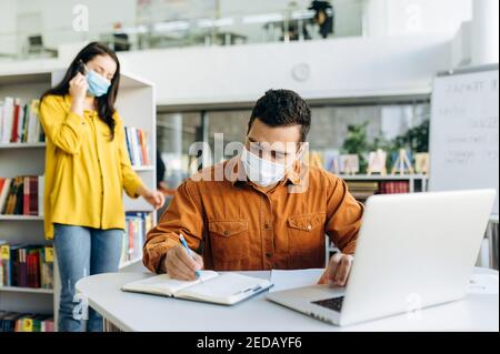 Successful male manager or student wearing protective medical mask, is working on a business project using a laptop, in the background a colleague talks on the phone in a protective mask Stock Photo
