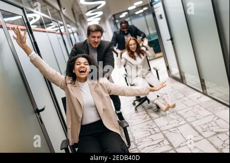 Group of multiracial business people in formal wear having fun at the workplace. Friendly coworkers make a race on office chairs. Young adult colleagues take a break from work Stock Photo