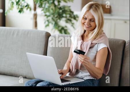 Senior happy woman shopping online. Joyful caucasian middle aged blonde woman holds and looks at the credit card, shopping online, online payment using laptop, sitting at living room Stock Photo