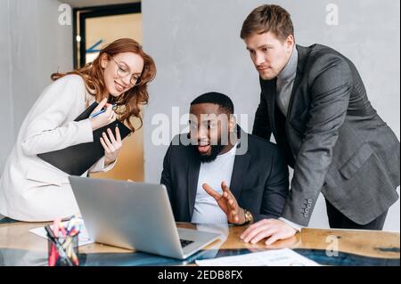 Happy multiracial business people in office using a laptop, look at the screen. Successful coworkers reading good news, get agreement for a project. Young adult employees working together, smiling Stock Photo