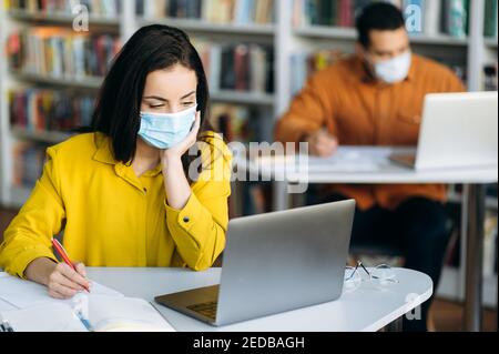 Education during quarantine. Students sit at a distance from each other dressed protective masks on their faces and studying using a laptop Stock Photo