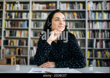 Portrait of a young satisfied caucasian woman wearing headset for working online, call center operator, consulting of client, or business mentor, looks away with smile Stock Photo