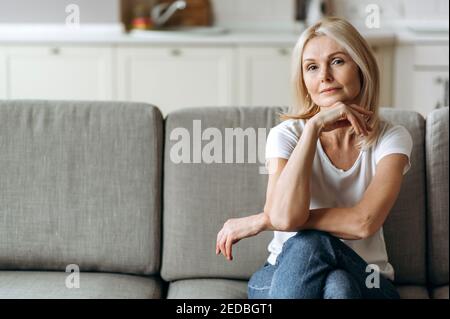 Portrait of charming calm caucasian senior blonde woman sits on couch at living room dressed in casual wear, looks at camera and smiling, weekend at home Stock Photo