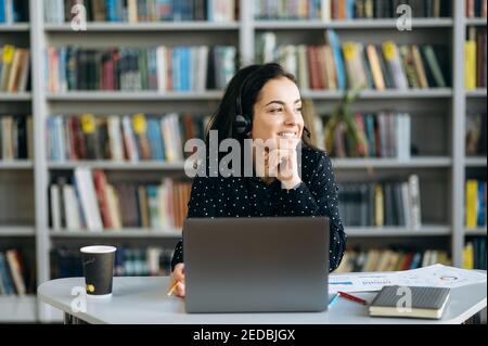 Pretty formal caucasian young woman, call center worker, student, or successful business tutor, in a headset is using a laptop while working online sitting at her work desk and looks away and smiling Stock Photo