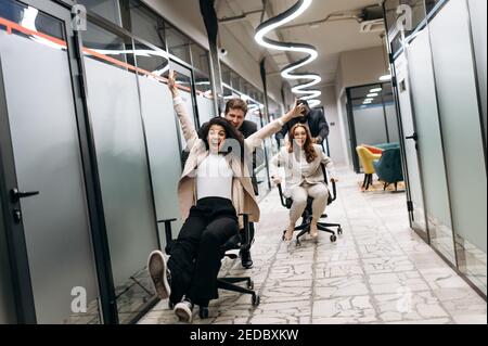 Young adult colleagues take a break from work. Friendly coworkers make a race on office chairs. Group of multiracial business people in formal wear having fun at the workplace Stock Photo