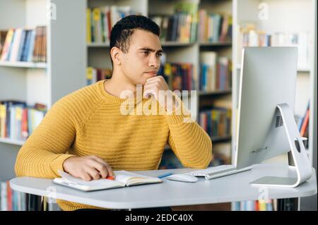 Hispanic man freelancer search ideas in the internet, using computer. Concentrated employee working on new startup. Focused business male sit at the desk in office Stock Photo