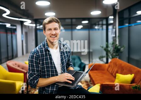 Caucasian business man standing in modern office, look at the camera, smiling. Happy male employee using laptop, working with new project or startup. Successful young adult manager at the office Stock Photo