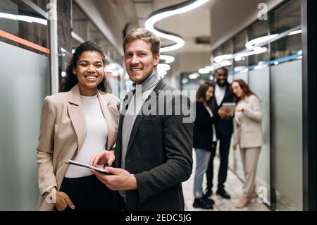Friendly business partners stand in modern office. Multiethnic business people in formal wear smiling, working together. Joyful male and female employee look at the camera, teamwork concept Stock Photo