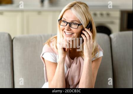 Attractive caucasian middle aged woman use smartphone, talking with friends or family. Senior female blonde in casual clothes sits on the couch with mobile phone, talks and smiling, communication Stock Photo
