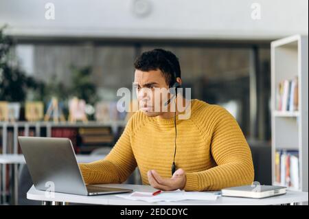 Confused male manager sits at the desk, using computer. Hispanic businessman have a problems with internet connection while online conference. Young adult employee worried about work issues Stock Photo