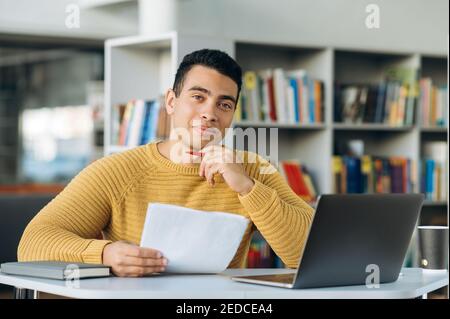 Portrait of young adult hispanic male freelancer or student is looking at the camera with smile. Attractive business man sits at the work desk, making notes, learning online Stock Photo