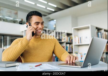 Joyful handsome businessman sitting in modern office, using smartphone, smiling. Happy hispanic male employee talking with friends or colleagues while working, communication concept Stock Photo