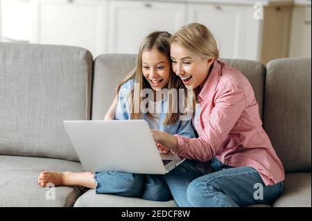 Leisure of mom and daughter. Happy caucasian mom and daughter are sitting on the couch, using a laptop, laughing, browsing the Internet, shopping online, got good news Stock Photo