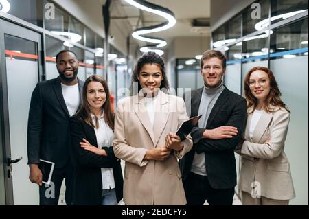 Portrait of successful influential business team in formal wear. Motivated female leader with employees of her company look at camera, smiling. Confident multiracial colleagues stand in modern office Stock Photo