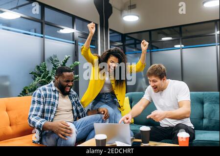 Overjoyed multiracial employees sitting in office, looking at the laptop, smiling. Successful business people happy about good deal. Cheerful students happy about good news Stock Photo