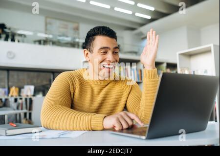 Handsome young adult man at online lesson or webinar training. Hispanic male student or employee sitting at the desk, using laptop and smiling. Studying remotely Stock Photo