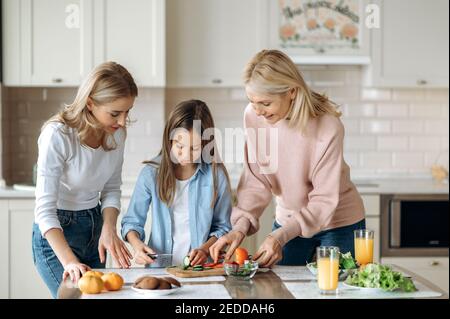 Three generations of women spend time together at the kitchen, thye teaches little girl to cut salad, smiling and happy to be together Stock Photo
