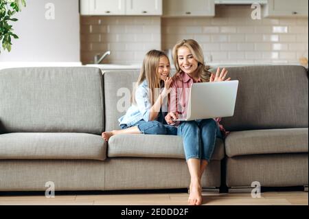 Video conference. Happy cheerful caucasian mom and daughter communicate online via video call using laptop while sitting at home in the living room, waving hands, greeting, smiling Stock Photo