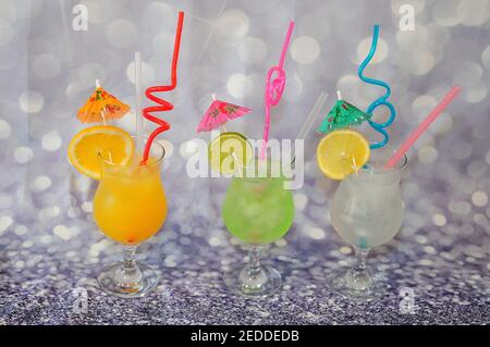 Three glasses with lime, lemon and orange juices with ice, straws and slices of citrus fruits, stand on a gray abstract background. Close-up. Stock Photo