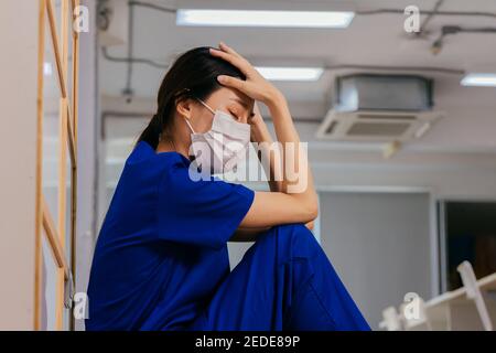 Young Asian woman nurse wearing uniform and surgical mask looking distraught and sad sitting on the floor while taking break after hard work in hospital Stock Photo