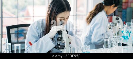 Young Asian female scientist doing research in laboratory wearing lab coat and gloves with colleague in background to develop antivirus vaccine Stock Photo
