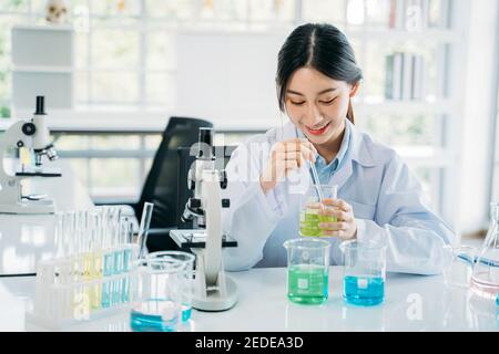 Young female Asian medical researcher working in pharmacy laboratory while mixing chemical in flask with tube and working on instruments like microscope and test tube Stock Photo