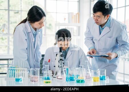 Group of young male and female Asian scientists wearing labcoat working together in lab using microscope to study and develop new antivirus vaccine Stock Photo