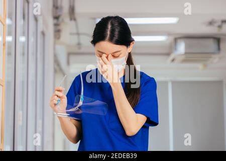 Young Asian nurse wearing uniform and rubbing eyes holding face shield in hand while wearing surgical mask looking distraught and sad while taking break after hard work in hospital Stock Photo