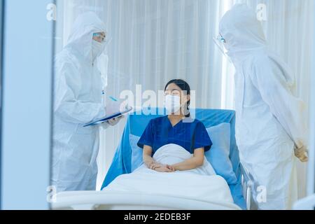 Covid-19 doctors in ppe suit with face shield and mask checking and treating young woman patient lying on bed suffering from coronavirus undergoing routine checkup Stock Photo