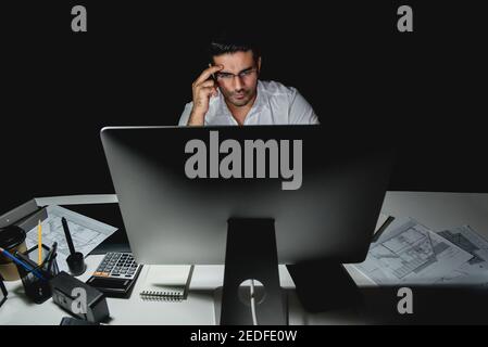 Serious Asian businessman or architect working late at night in the office looking at computer screen and thinking Stock Photo