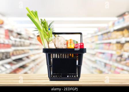 Food and groceries in black shopping basket on wood table with blurred suppermarket aisle in background Stock Photo