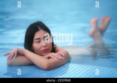 Glamorous brunette woman lies in thermal water in pool at spa and relaxes. Soft selective focus on model's eyes Stock Photo