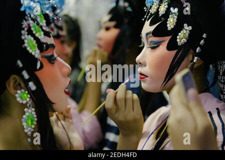 A profile closeup of a young beautiful Chinese opera player in a magnificent fairy costume, while her friend brushing her lips with a lipstick brush. Stock Photo